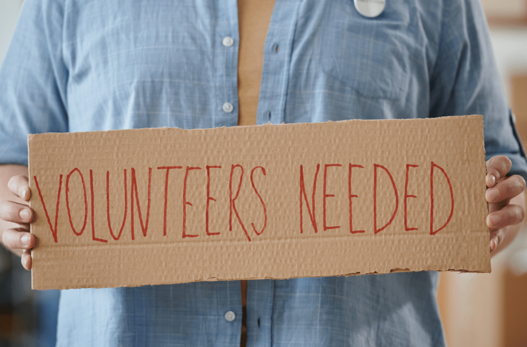 Mensch hält ein Papp-Plakat hoch, auf dem steht: Volunteers neede - Ehrenamtliche gebraucht