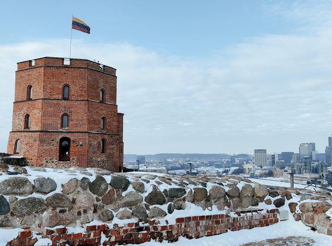 Hinter einer Steinmauer steht ein kleiner Backstein-Turm, auf dem eine Litauen-Flagge weht. Im Hintergrund sieht man eine Großstadt