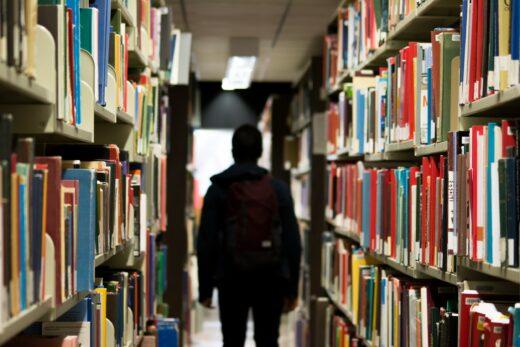 Eine junge Person steht zwischen wandhohen Bücherregalen einer Bibliothek.