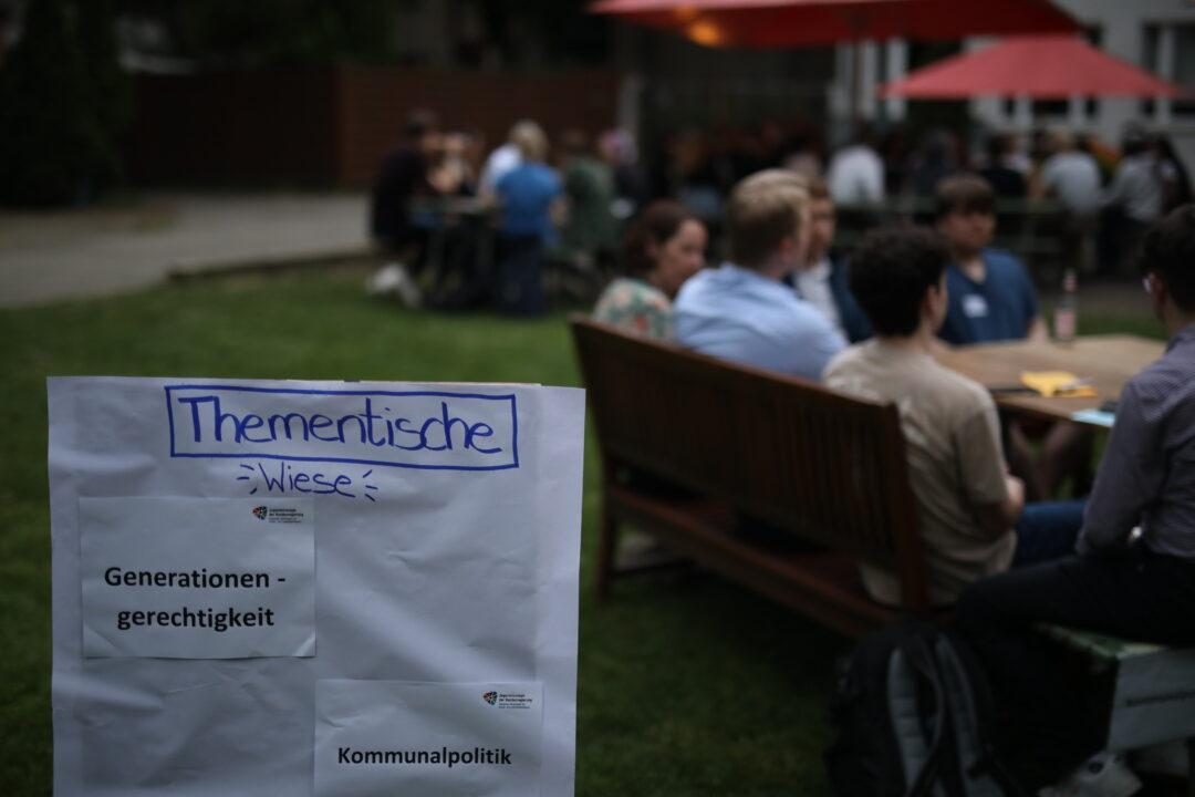 Im Hintergrund sitzen junge Menschen an einem Tisch. Vorne ist ein Aufsteller mit der Aufschrift Thementisch zu sehen.