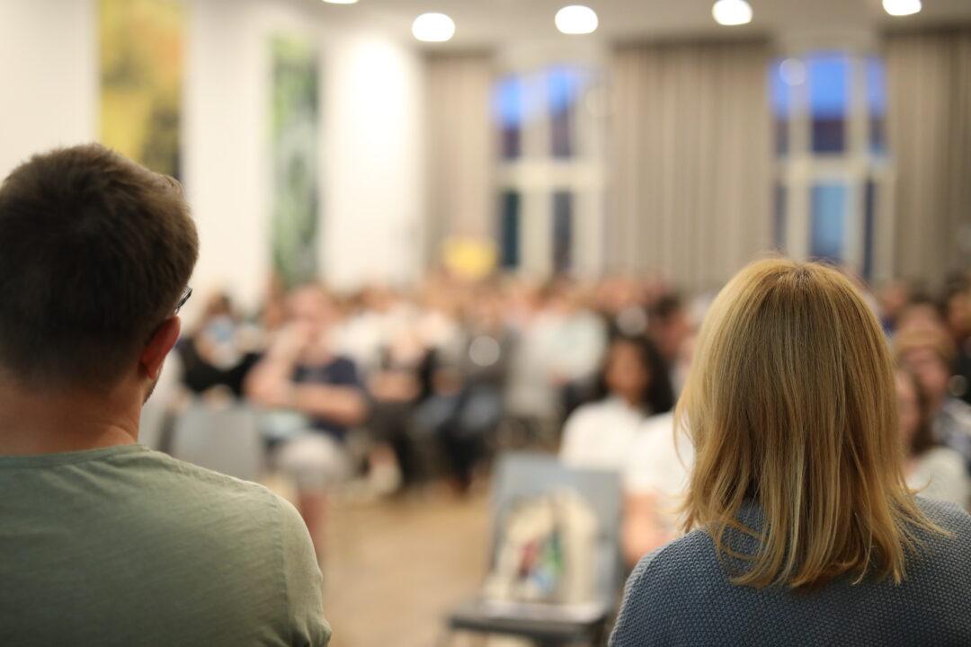 Zwei Menschen sitzen auf einem Podium. Sie sind von hinten zu sehen.